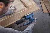 安全装備を身に着けた人がオービタルサンダーで木製のドアを研磨しています。.