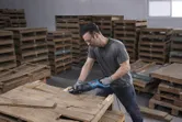 安全装置を装着した人がレシプロソーで木製パレットを切断しています。