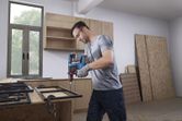安全装置を装着した人が木製の作業台の上で金属フレームに穴を開けています。