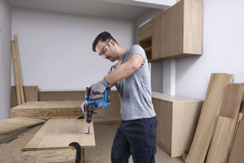 安全装置を装着した人が、コードレス回転ハンマーを使用して木材に穴を開けています。.