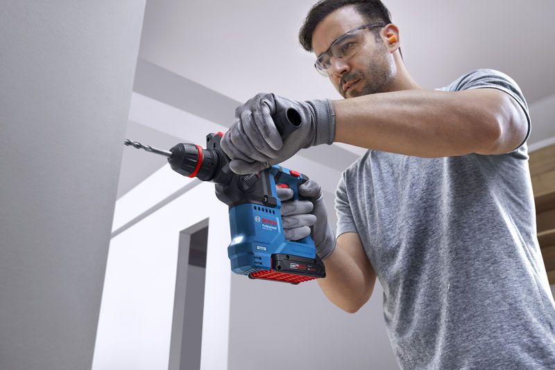 安全装置を装着した人が、コードレス回転ハンマーを使用してコンクリートの壁に穴を開けています。.