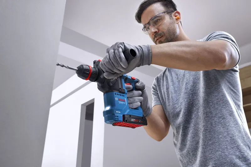 安全装置を装着した人が、コードレス回転ハンマーを使用してコンクリートの壁に穴を開けています。.