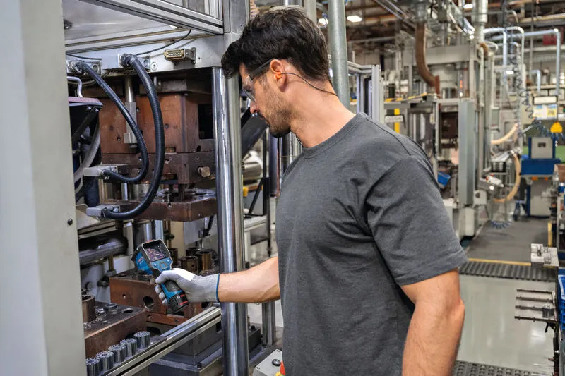 工場内で安全装備を身に着けた人がサーモカメラを使って機械を検査しています。.