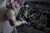 安全装備を身に着けた人が、コードレスラチェットレンチを使用して自動車のエンジンを作業しています。
