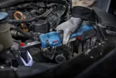 安全装備を身に着けた人が、コードレスラチェットレンチを使用して自動車エンジンのボルトを締めています。
