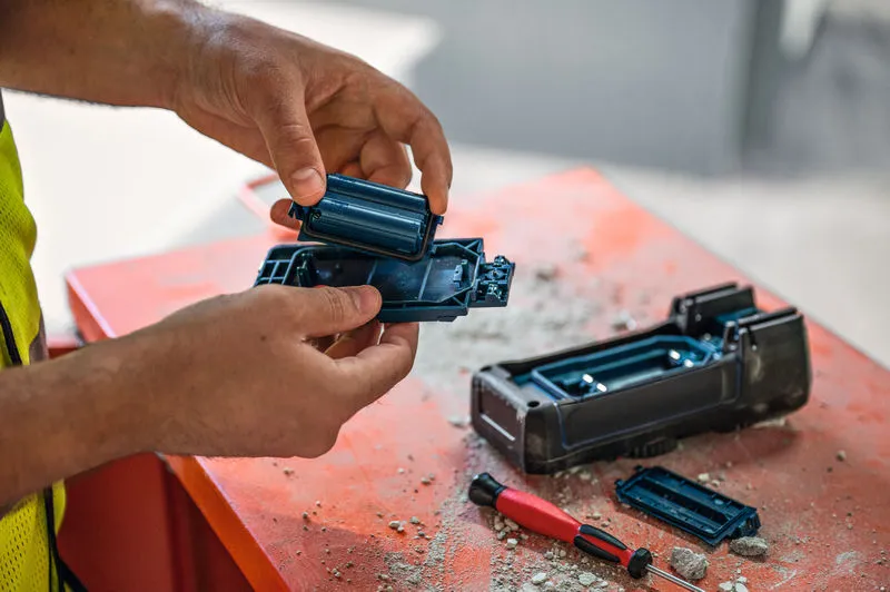 安全装置を身に着けた人が、埃っぽい作業台の上で電動工具の電池を交換しています。.
