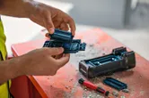 安全装置を身に着けた人が、埃っぽい作業台の上で電動工具の電池を交換しています。