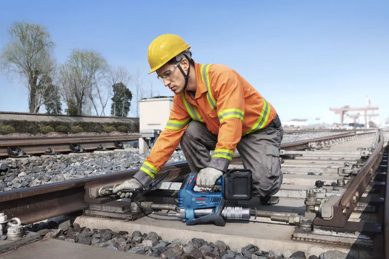 安全装備を身に着けた作業員が、電動工具を使って鉄道線路のボルトを締めている。