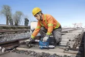 安全装備を身に着けた作業員が、電動工具を使って鉄道線路のボルトを締めている。