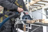 安全装備を身に着けた人が足場の上でコードレス丸鋸を使って木材を切っています。.