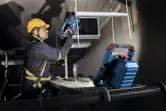 安全装置を装着した人が、明るい現場照明の下でコードレス電動工具を使用しています。.