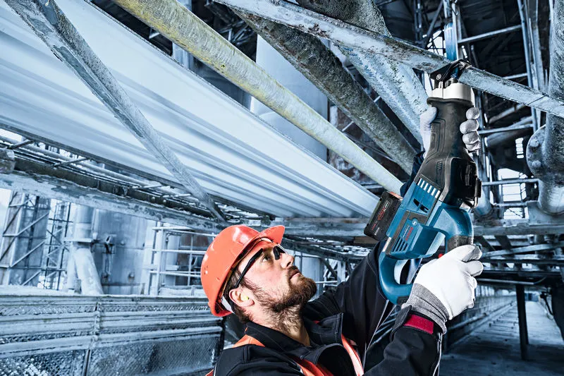 安全装置を装着した人が往復鋸を使用して頭上の金属パイプを切断しています。.