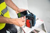 安全装備を身に着けた人が、はしごの上で携帯用電源を使って携帯電話を充電している。