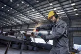 安全装備を身に着けた人が工場の作業場でアングルグラインダーを使って金属パイプを研磨しています。.