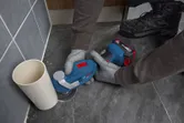 安全装置を装着した人が多機能工具を使ってパイプの横の床タイルを切断しています。