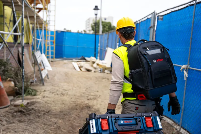 安全装備を身に着けた人が、建設現場で作業員用のバックパックとキャリングケースを携行している。