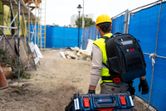 安全装備を身に着けた人が、建設現場で作業員用のバックパックとキャリングケースを携行している。