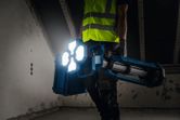 安全装備を身に着けた人が薄暗い部屋でコードレスの現場用ライトを運んでいます。