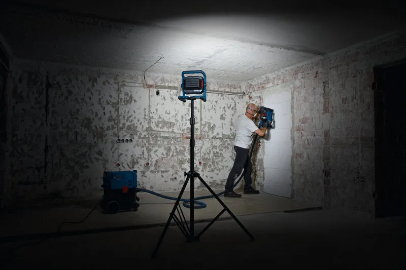 安全装備を着用した人が作業灯の下で壁の穴あけ工具を操作しています。