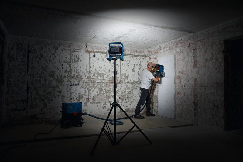 安全装備を着用した人が作業灯の下で壁の穴あけ工具を操作しています。