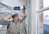 安全装備を身に着けた人が窓の近くの壁枠にドリルで穴を開けています。