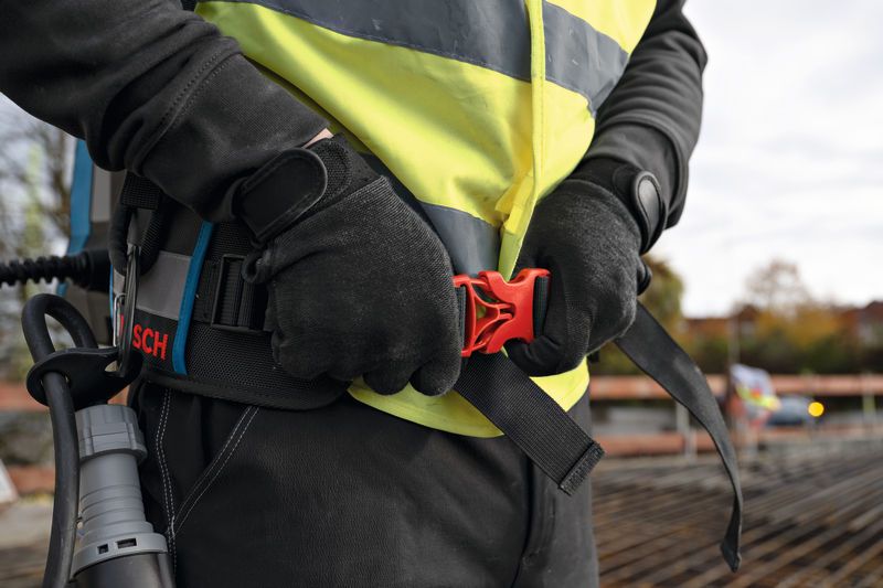 安全装備を身に着けた人が、建設現場でバックパックに取り付けられたベルトを調整している。