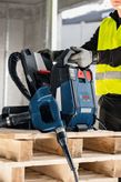 安全装備を身に着けた人が木製パレットの上で集塵機とドリルを操作しています。