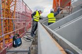 安全装備を身に着けた2人が建設用具とコンクリート型枠を使って足場で作業しています。