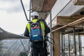 安全装備を身に着けた人が、屋外の足場の上に立ち、青い背負い式の掃除機システムを手にしている。