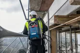 安全装備を身に着けた人が、屋外の足場の上に立ち、青い背負い式の掃除機システムを手にしている。