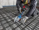 安全装置を装着した人が、建設現場でコードレス工具を使って鉄筋を切断しています。