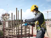 安全装備を身に着けた人が、コードレス工具を使って建設現場で鉄筋を切断しています。