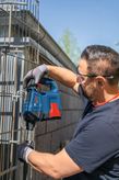 安全装備を身に着けた人が、電動工具を使って金属製のフェンスに穴を開けている。