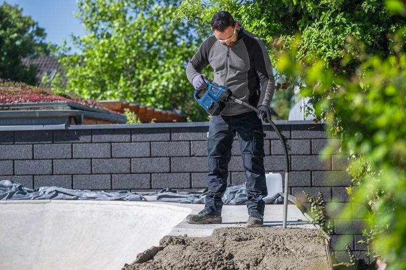 安全装備を身に着けた人が、屋外で電動工具を使ってコンクリートを締め固めている。