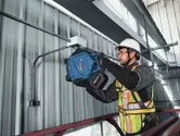 安全装備を身に着けた人が、工業用建物の金属製の壁にドリルで穴を開けている。