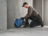 安全装備を身に着けた人が、電動工具を使ってコンクリートに溝を掘っている。