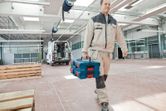 安全装備を身に着けた人が、広々とした作業場で積み重ねられた工具箱を運んでいます。